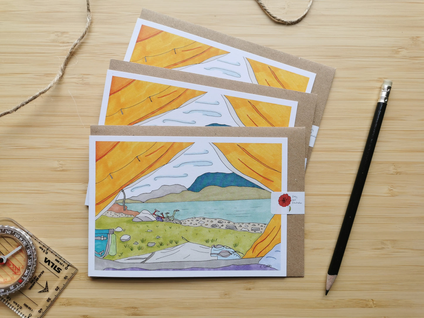 Set of greeting cards with scenic illustrations looking out of a tent with a view of a bike and a loch, on a wooden surface with a pencil and ruler.