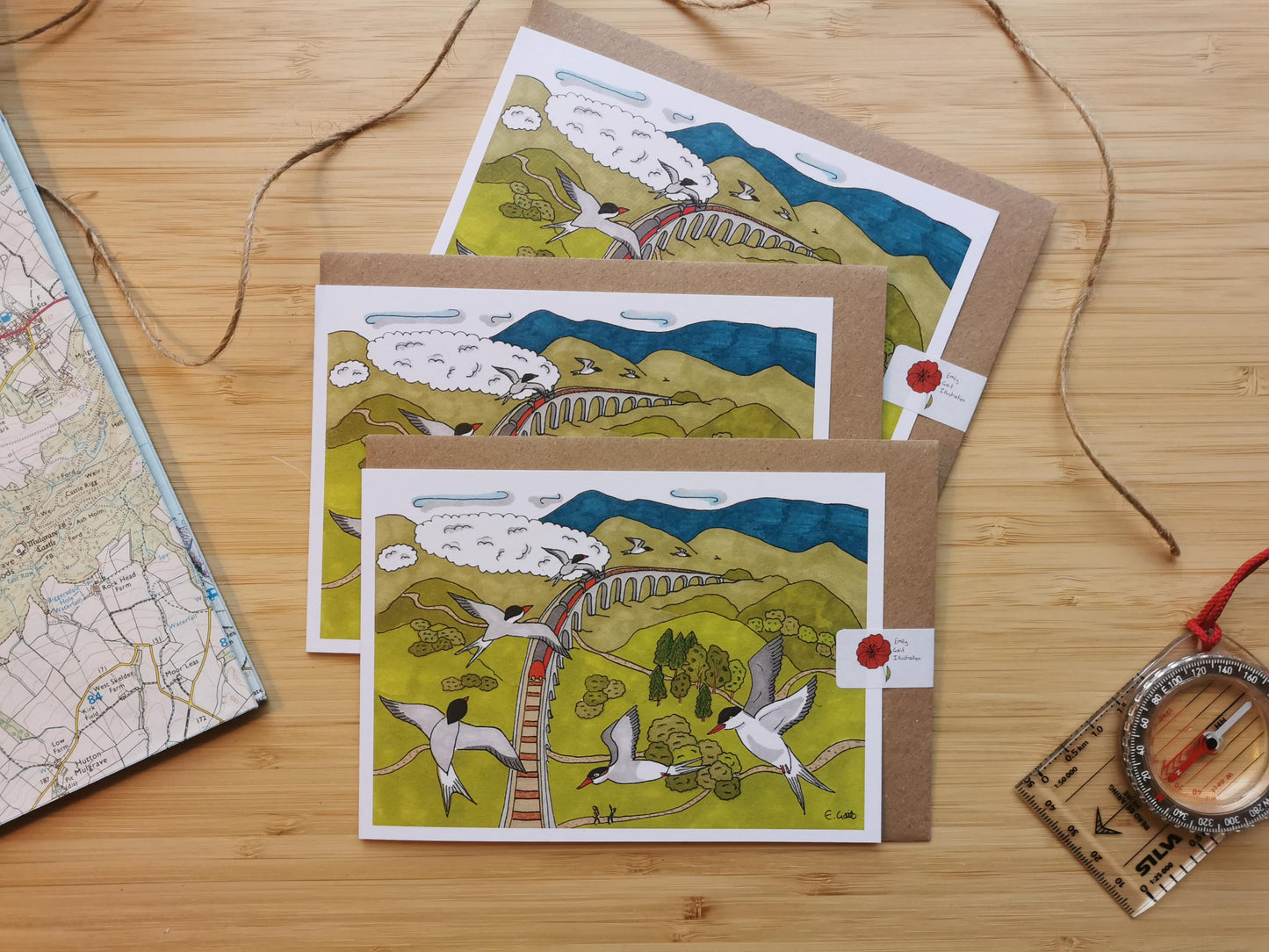 Three illustrated cards of artctic terns and a steam train going over a viaduct in the Scottish Highlands, on a wooden surface with maps and a compass.