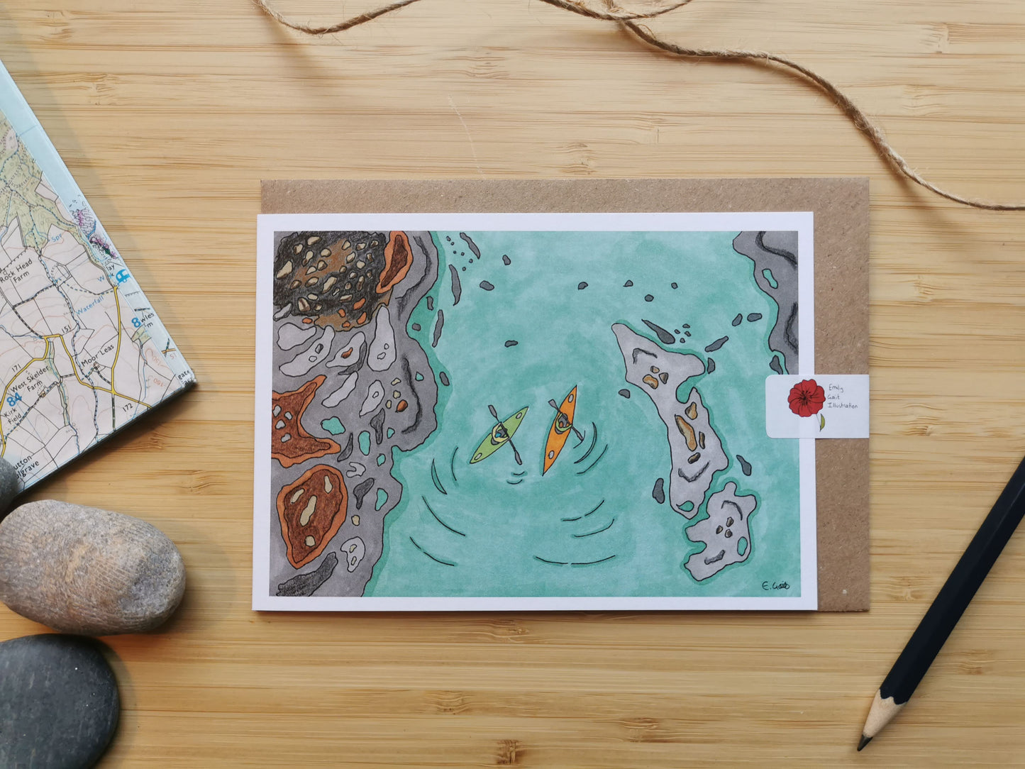 Illustrated card with kayaks on water, placed on a wooden surface with rocks and a map.