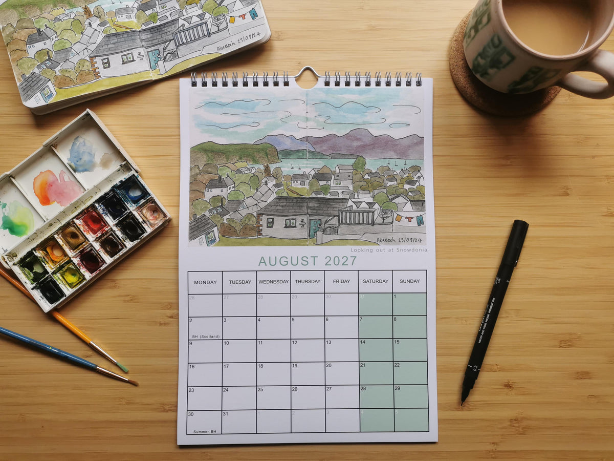 A spiral-bound August 2027 desk calendar featuring a hand-painted village landscape, styled on a wooden tabletop with a watercolor palette, paintbrushes, a black pen, and a ceramic mug.