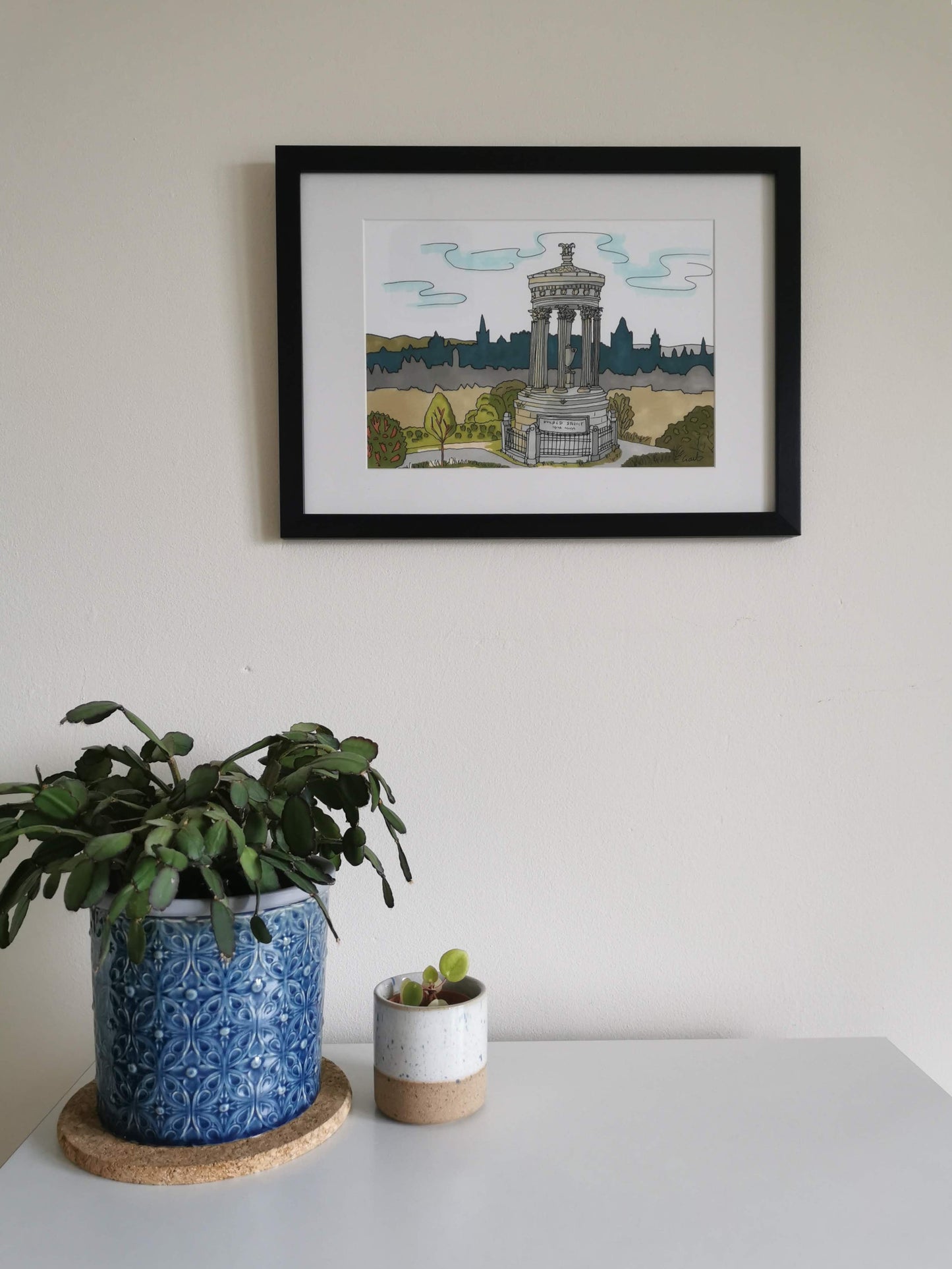 Framed artwork of an illustration from the top of Calton Hill in Edinburgh, on a wall above two potted plants.