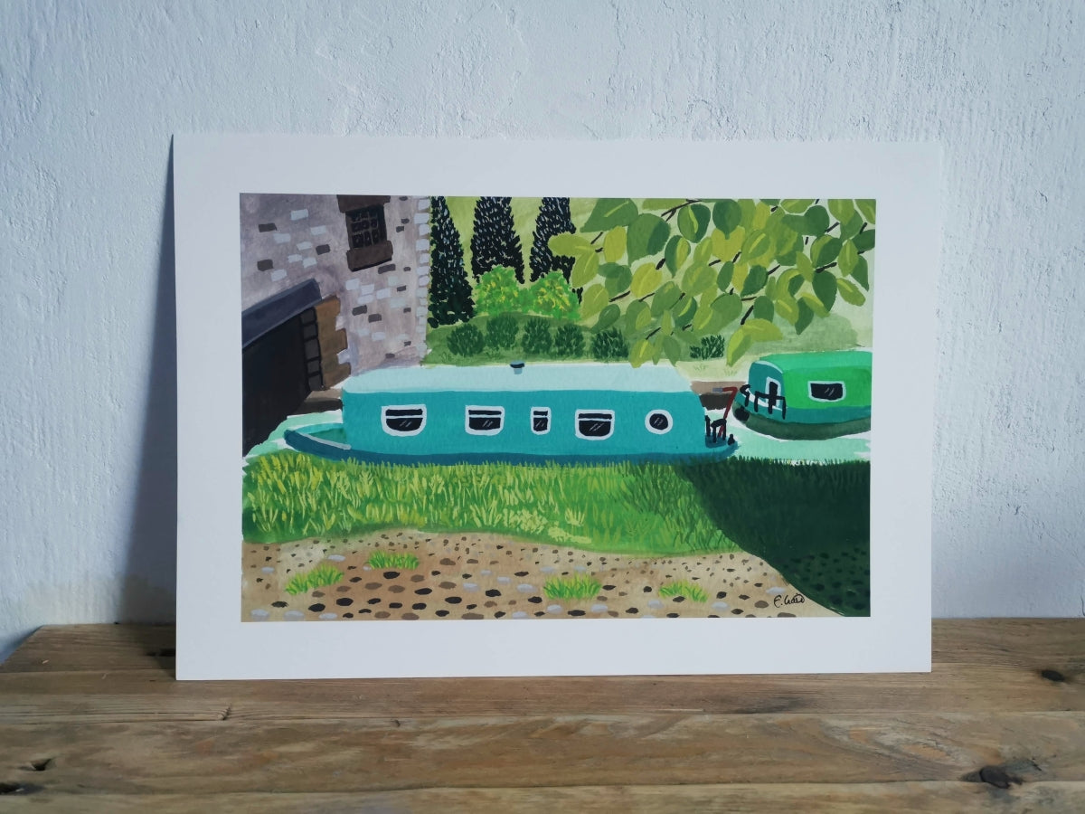 Framed artwork of a teal houseboat with a green landscape on a wooden surface.