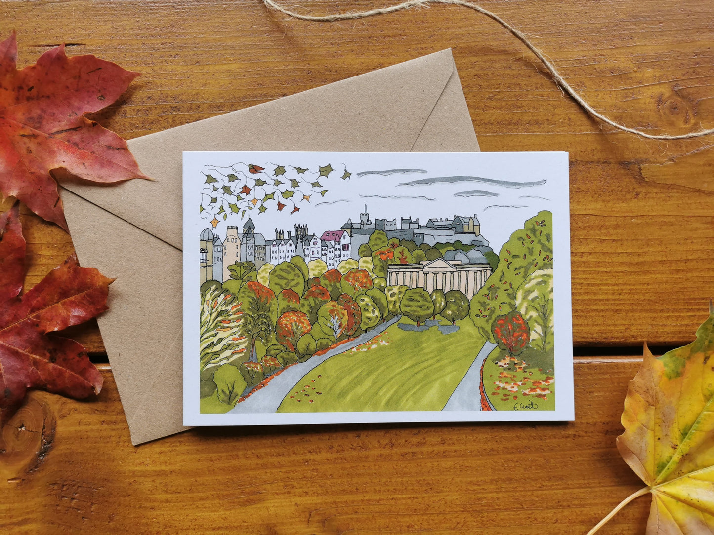A card featuring a colourful autumnal pen and ink illustration of Edinburgh Castle with Princes Street Gardens  and autumnal leaves in the foreground. 