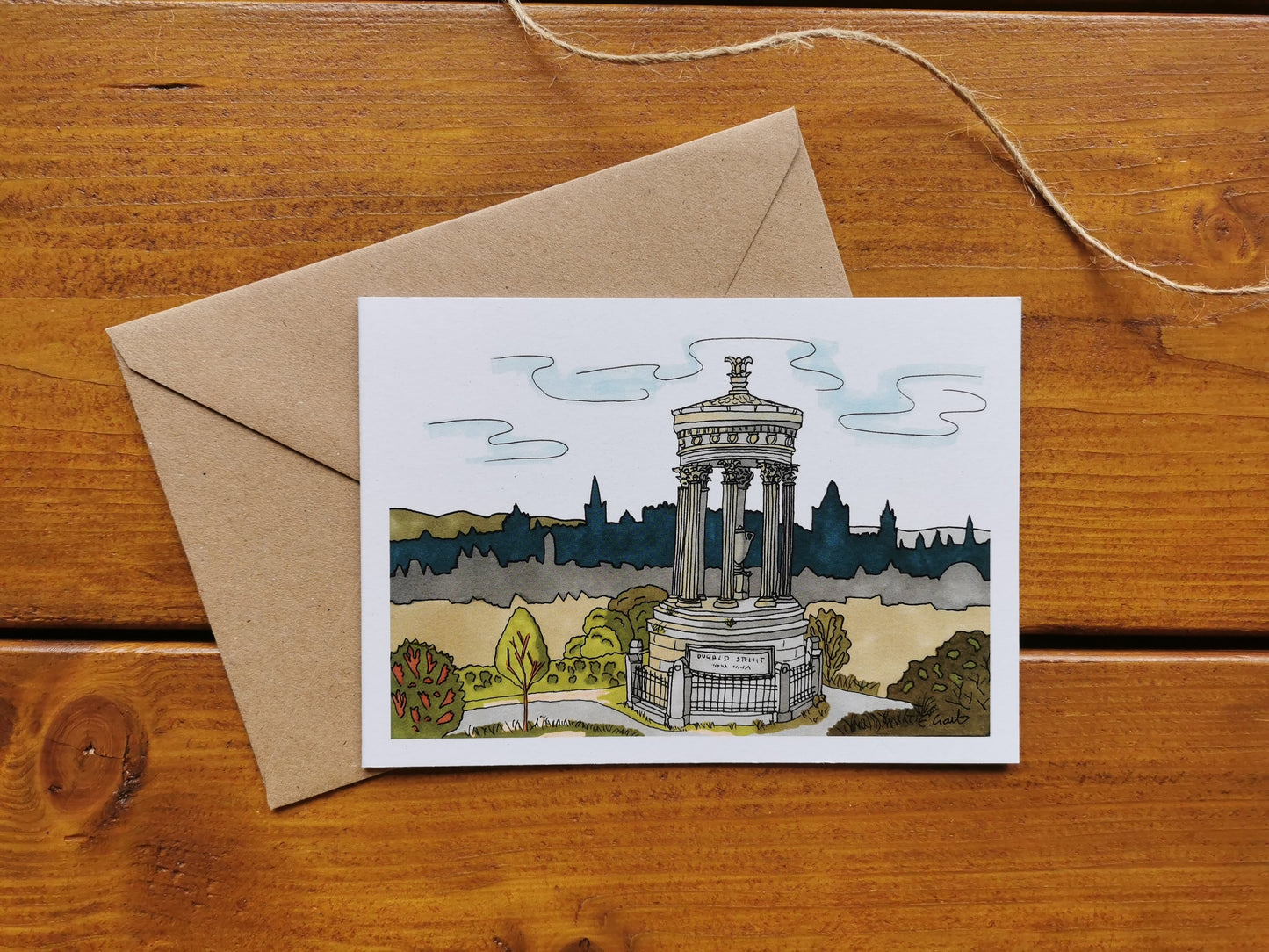 A greeting card featuring a drawing of Calton Hill and the Nelson Monument with Edinburgh in the background, placed on a wooden surface with a brown kraft envelope.