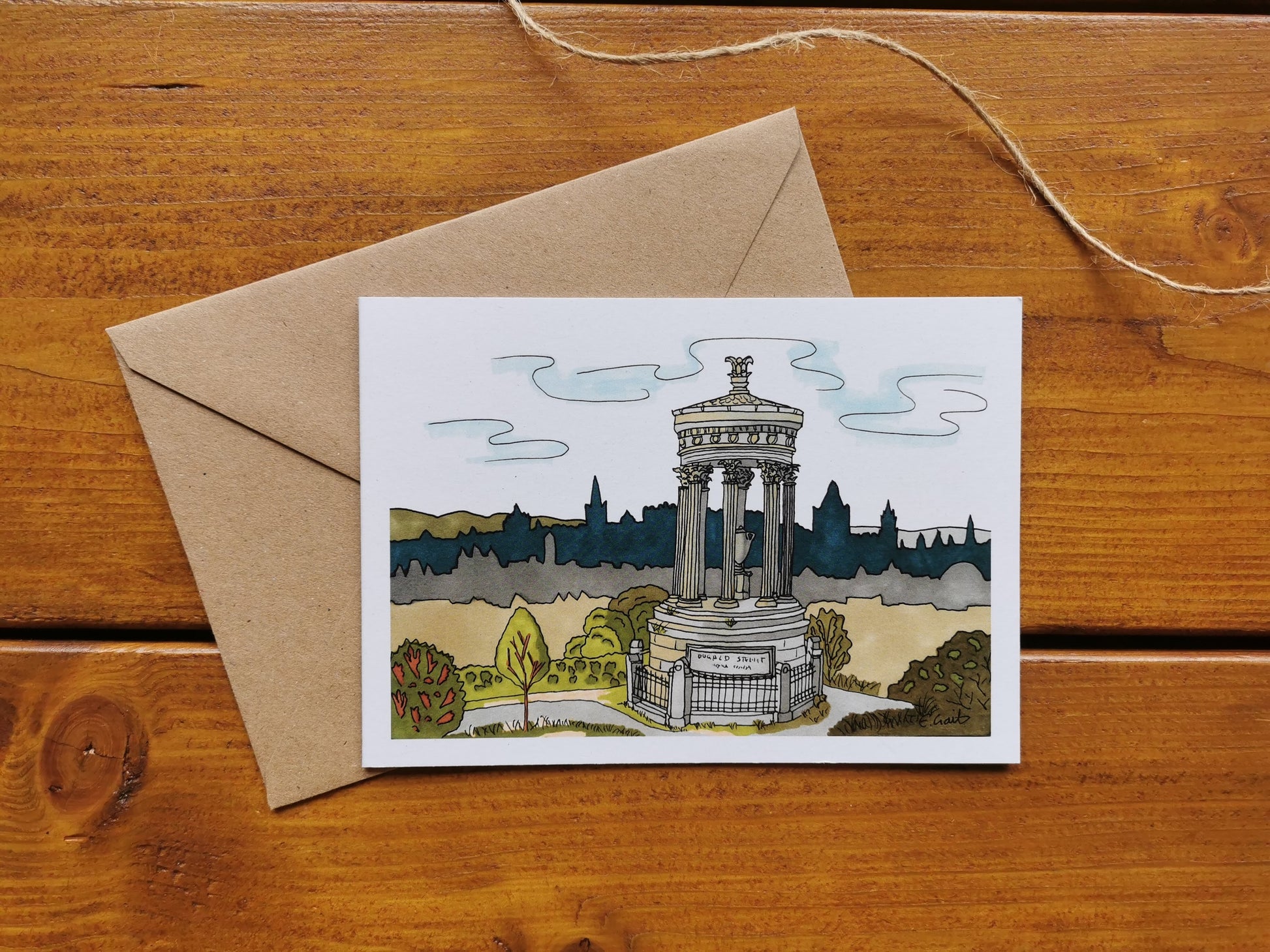A greeting card featuring a drawing of Calton Hill and the Nelson Monument with Edinburgh in the background, placed on a wooden surface with a brown kraft envelope.