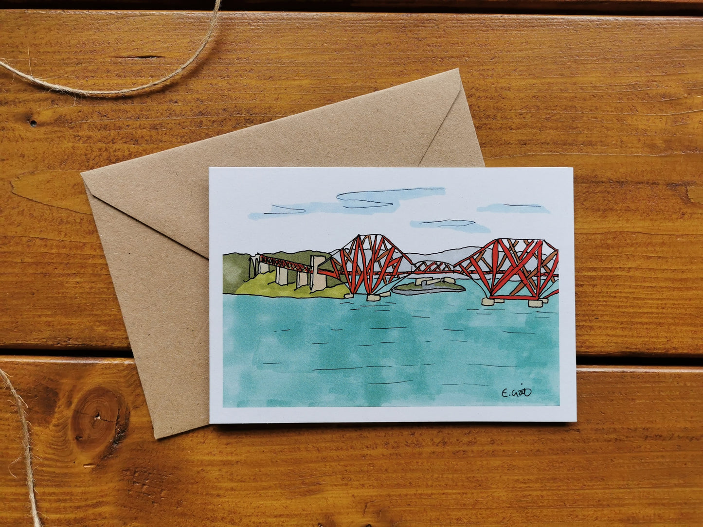 A greeting card with an illustration of the Forth Rail Bridge in Scotland, accompanied by a brown kraft envelope on a wooden surface. The card is blank inside.