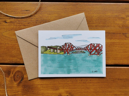 A greeting card with an illustration of the Forth Rail Bridge in Scotland, accompanied by a brown kraft envelope on a wooden surface. The card is blank inside.