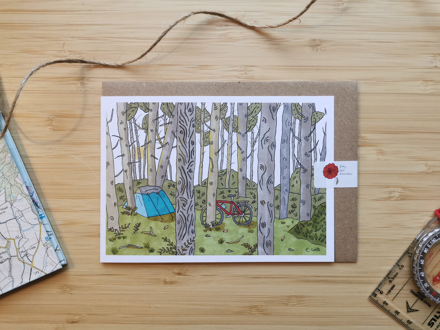 Card with illustration of a forest scene with a tent and bike, on a wooden surface.