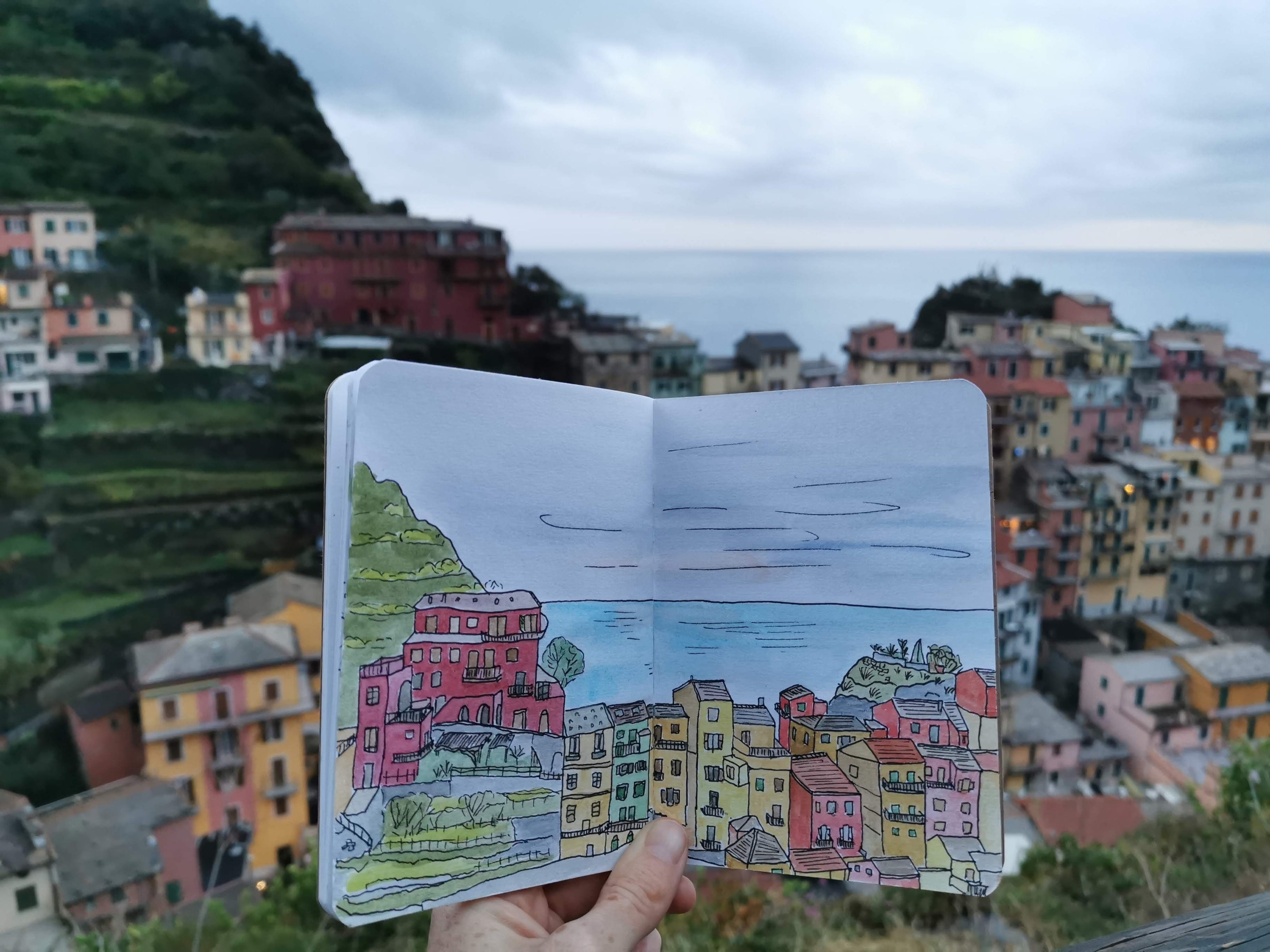 Photo of a painting in a sketchbook of colourful buildings in Italy, held up against a real landscape of colourful buildings. The sea is in the background.
