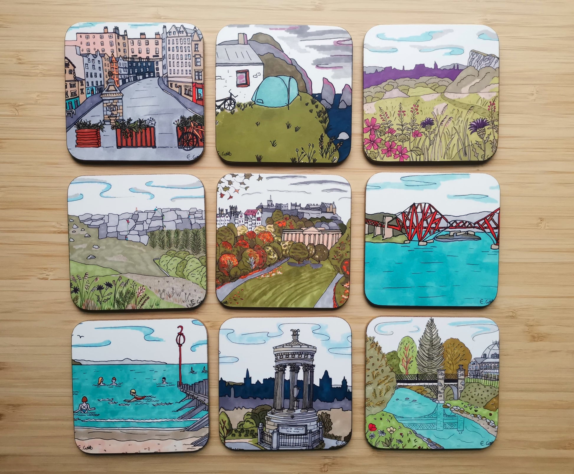 Set of 9 illustrated coasters on a wooden surface. The designs include Victoria Street in Edinburgh, Camping next to the sea, Arthur's Seat, Climbing on Stanage Edge, an Autumn park scene in Edinburgh, Forth Rail Bridge, people swimming on the beach, a view from Calton Hill with Edinburh in the background and a park scene with Buxton Pavilion in the background.