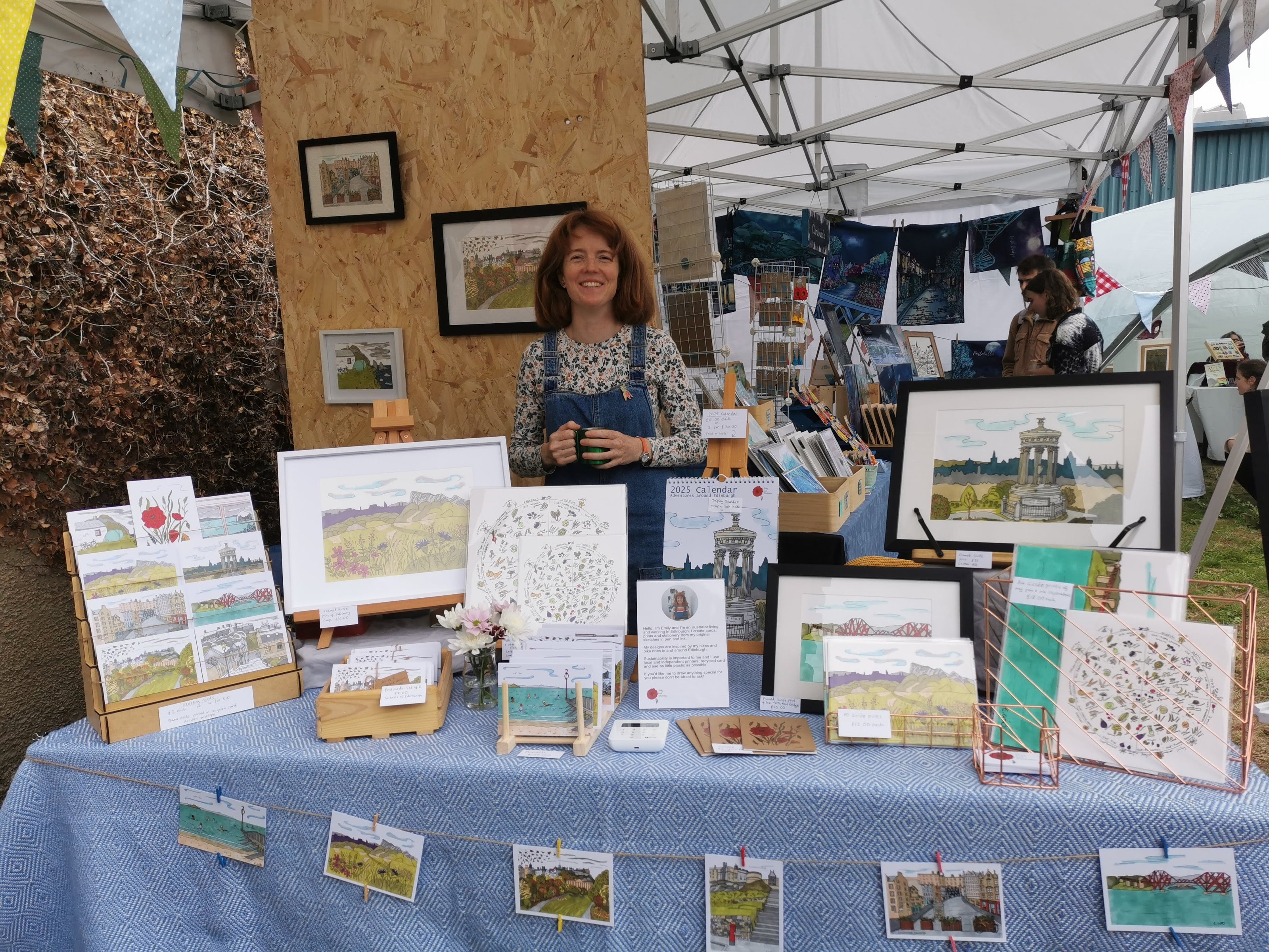 Photo of a woman behind a table full of artwork, greeting cards and notebooks.
