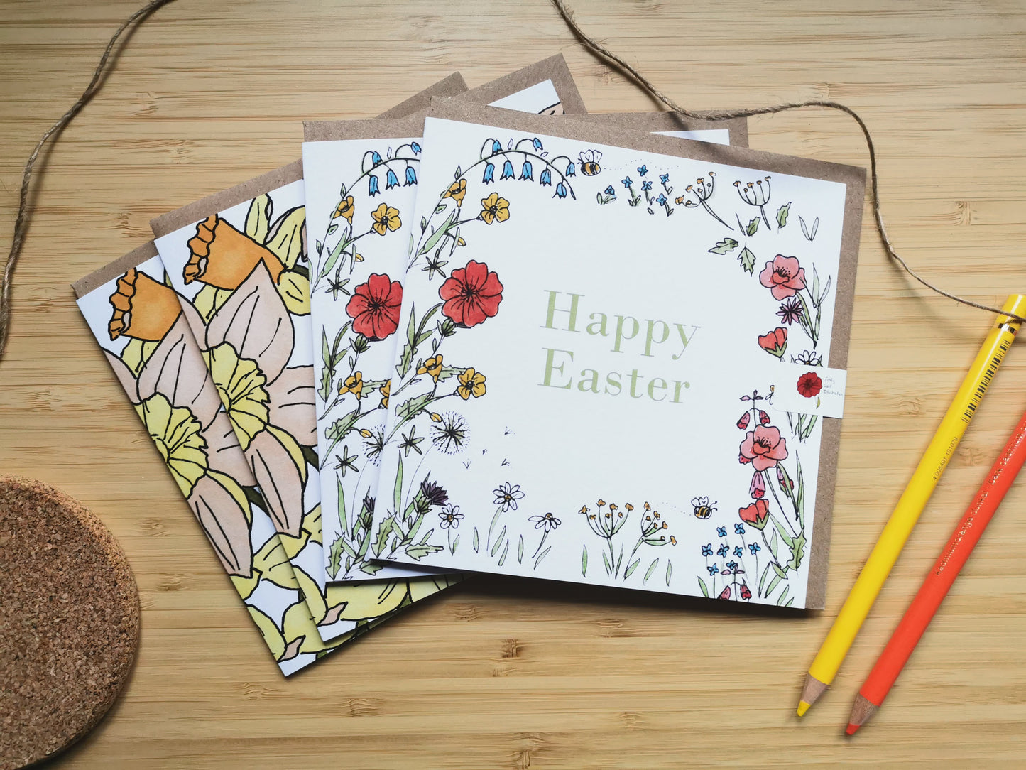 Two easter greeting cards with wild flowers and daffodil illustrations on a wooden surface with pencils.