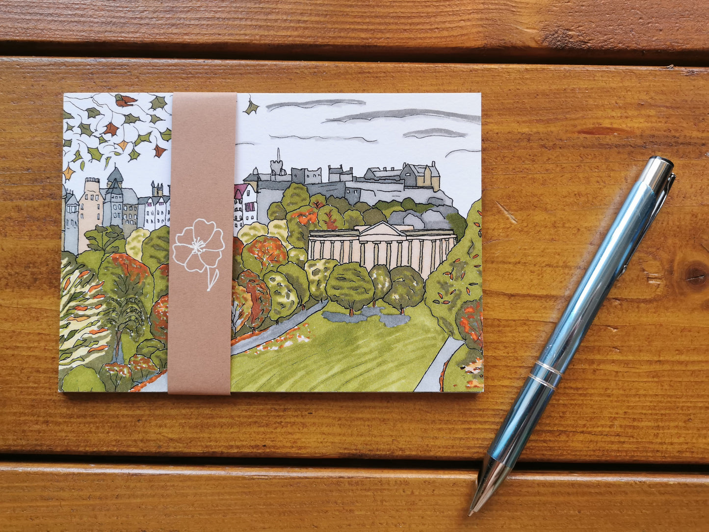 A Postcard set of Edinburgh illustrations wrapped up in a paper sleeve, on a wooden table with a pen next to it.