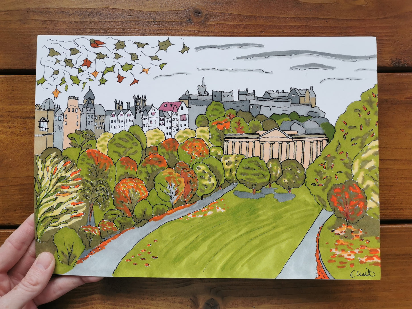 A framed Giclée print of 'Autumn in Edinburgh' displayed on a wooden easel, featuring an illustration of a park with trees and a castle in the background, next to a pumpkin and a green plant.
