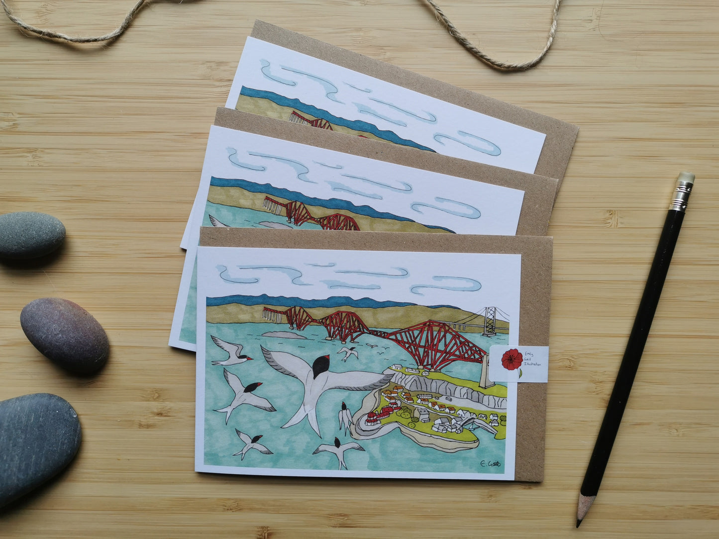 Set of three illustrated greeting cards on a wooden surface with a pencil and stones.