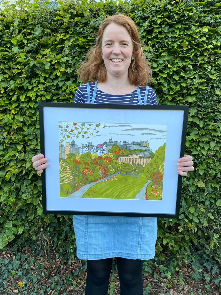 Photo of a woman holding a framed illustration of an autumn landscape with Edinburgh Castle in the background and autumn leaves and colours.