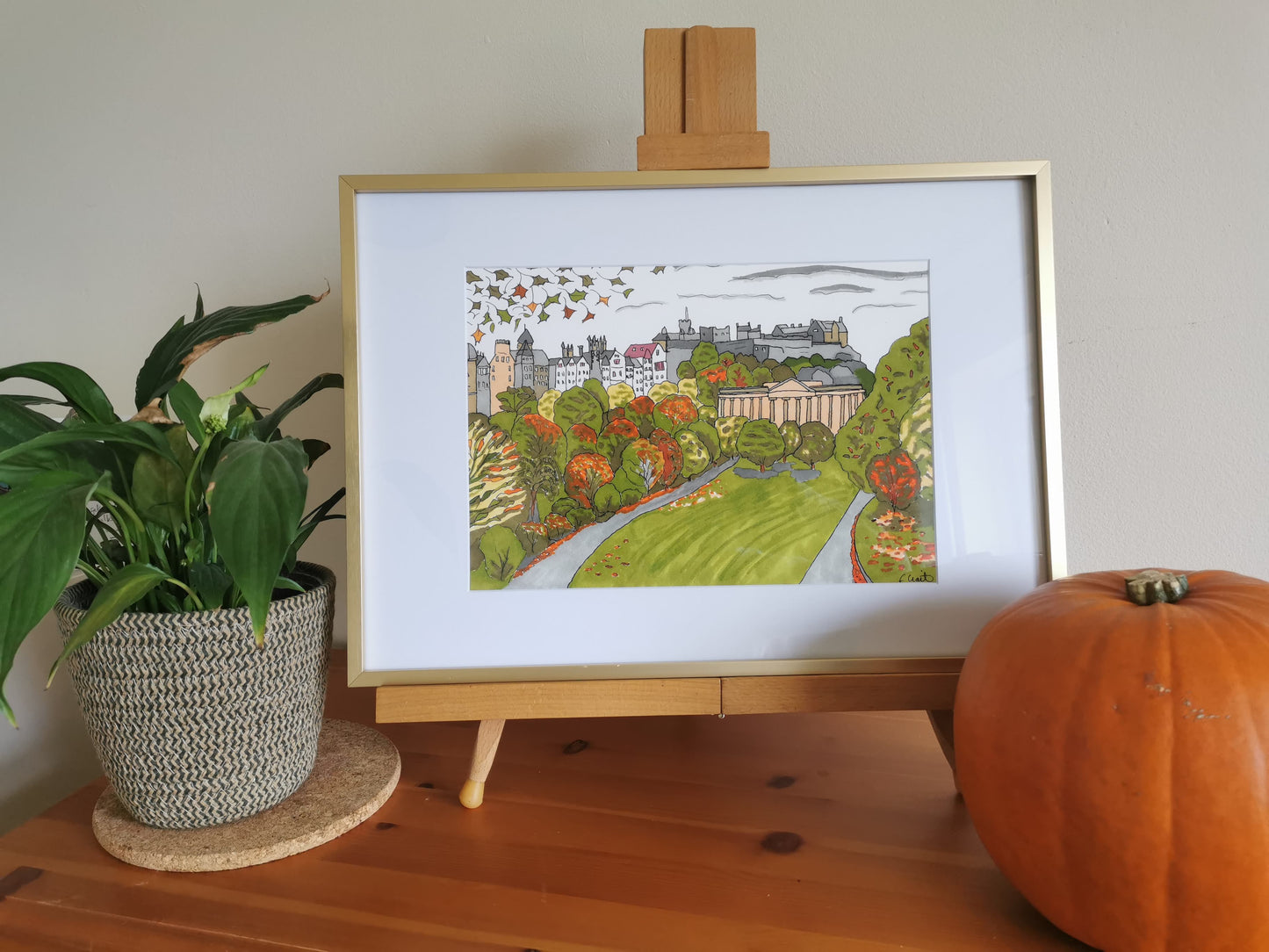 A framed Giclée print of 'Autumn in Edinburgh' displayed on a wooden easel, featuring an illustration of a park with trees and a castle in the background, next to a pumpkin and a green plant.