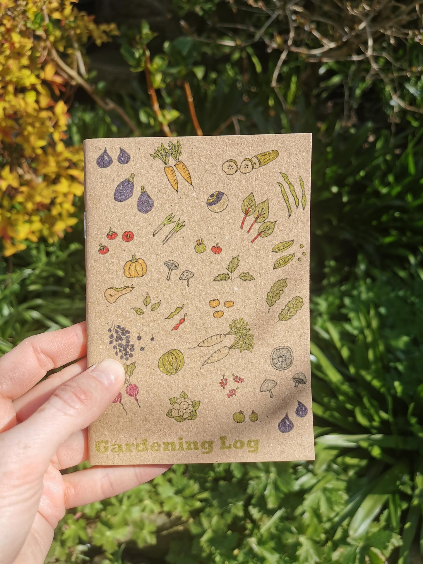 A hand holding a small A6 gardening log notebook with a pen and ink illustration of various fruits and vegetables on its cover.