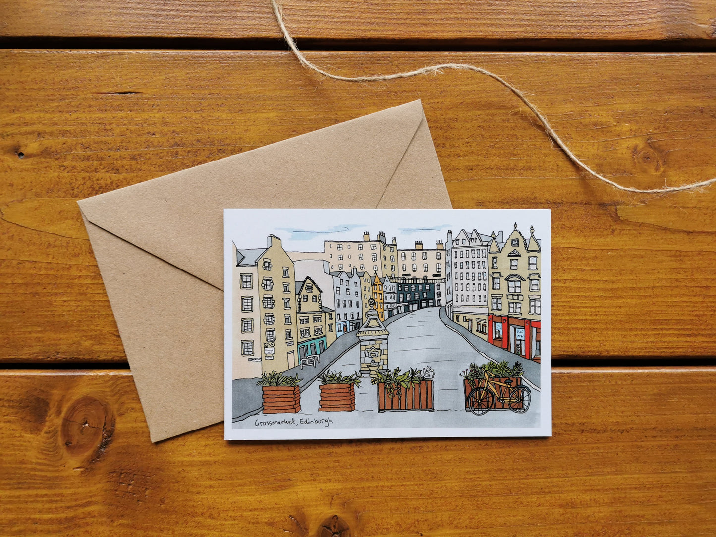 A greeting card depicting a illustration of Victoria Street, Edinburgh, laid on a wooden surface with its brown kraft envelope, tied with a string.