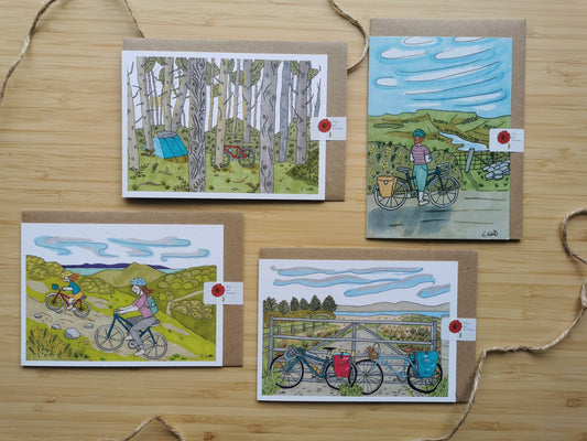Four illustrated greeting cards on a wooden surface with cyclists and nature scenes.