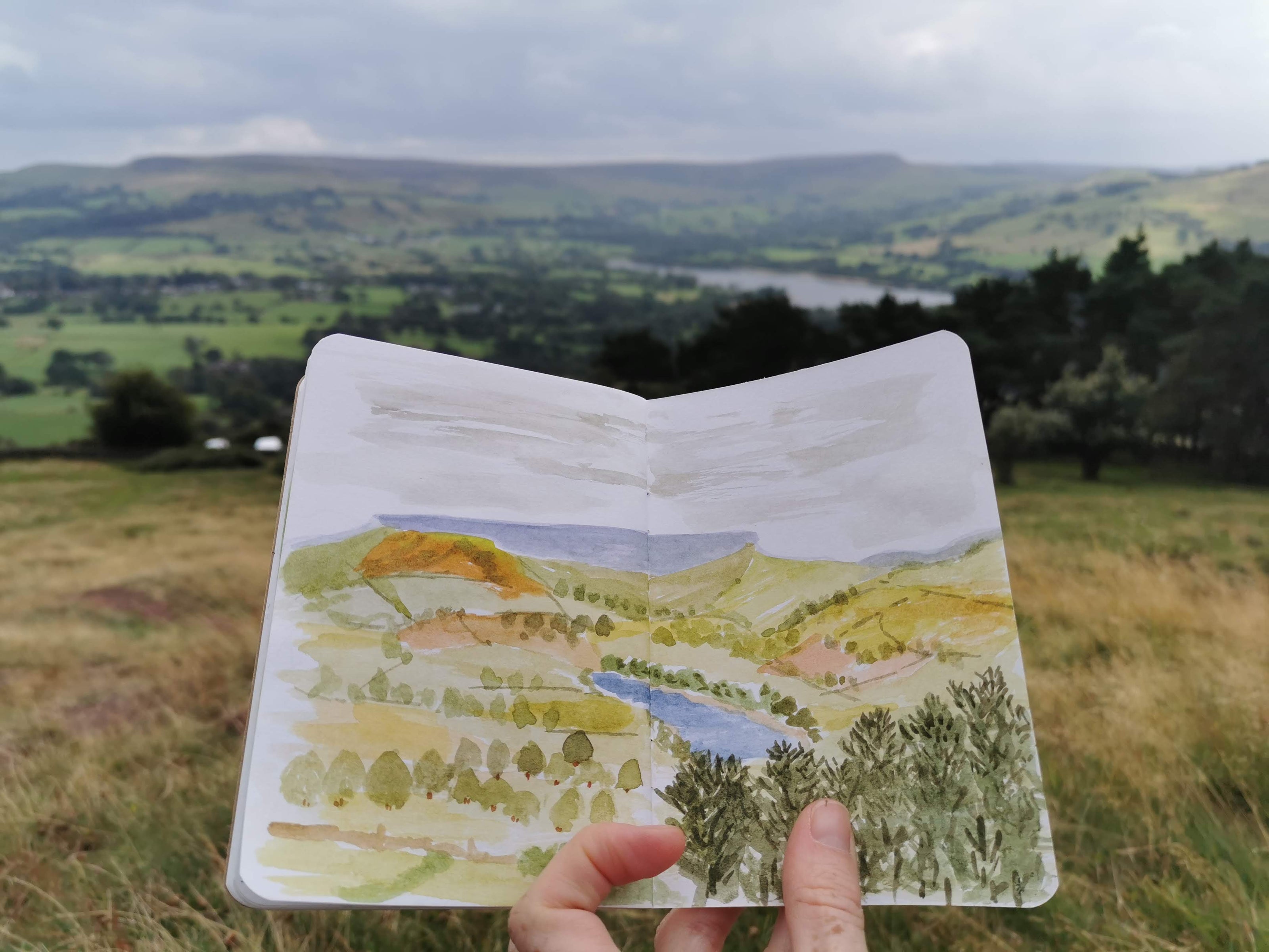 painting in a sketchbook of the view in situ - a landscape with a lake in the middle surrounded by trees and hills.