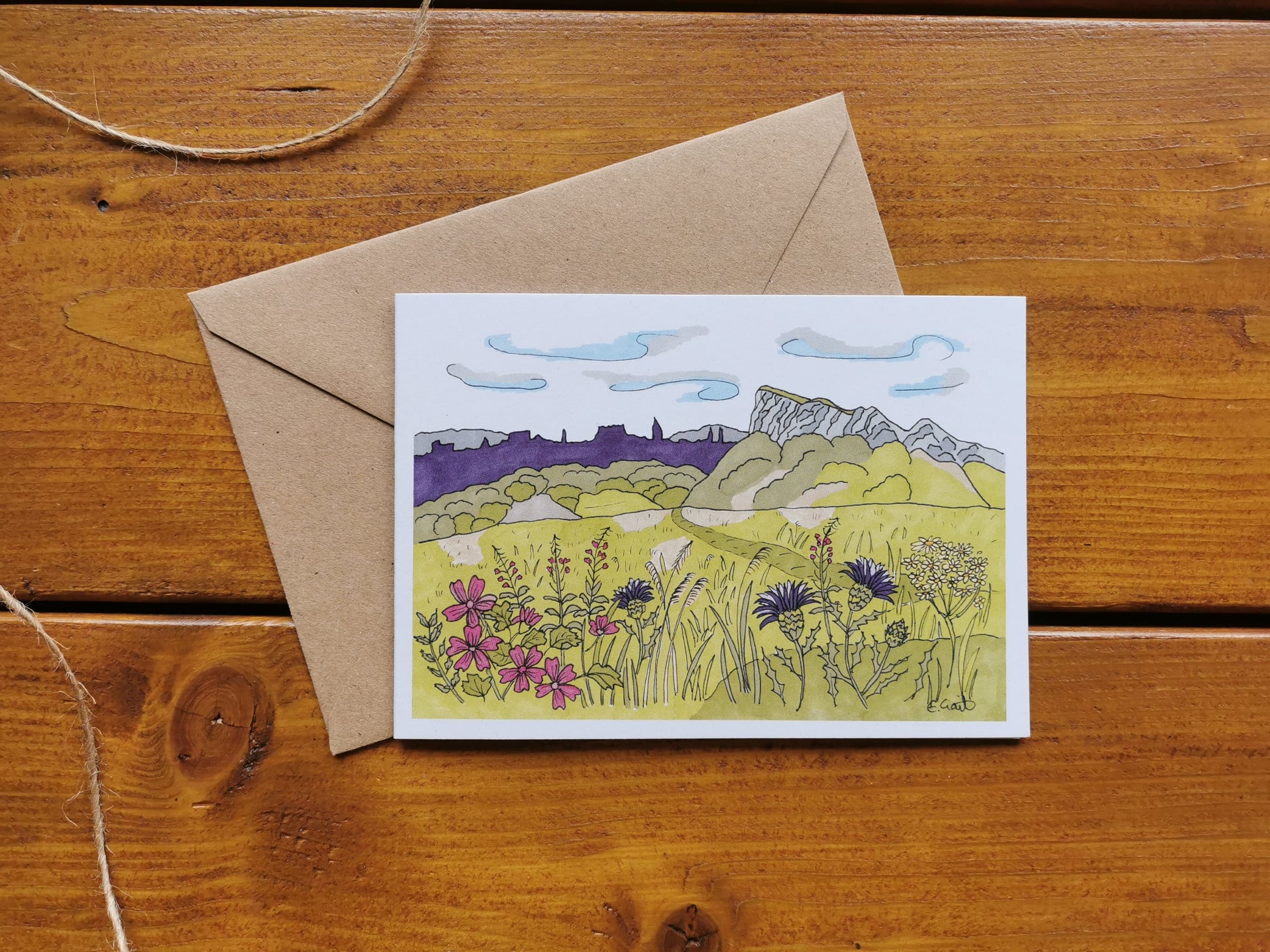 A greeting card featuring a illustration of Salisbury Crags with a brown kraft envelope on a wooden background.