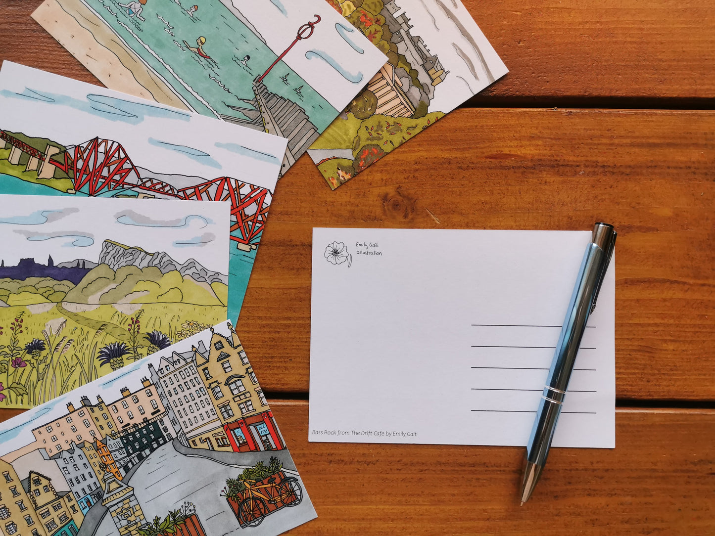 A set of six illustrated postcards depicting various scenes of Edinburgh, displayed on a wooden table with a pen.