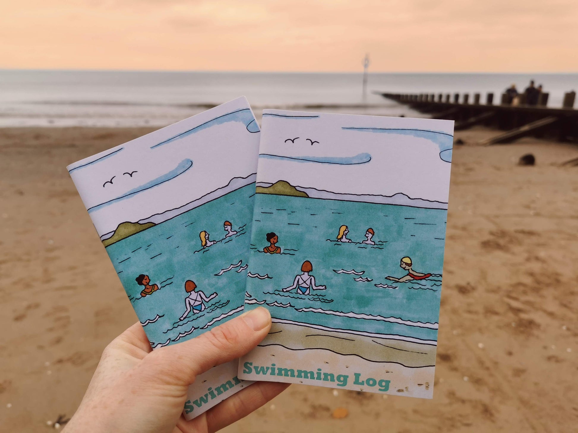 A hand holding an A6 size swimming log notebook with a illustration of people swimming in the sea on the cover, at a beach.