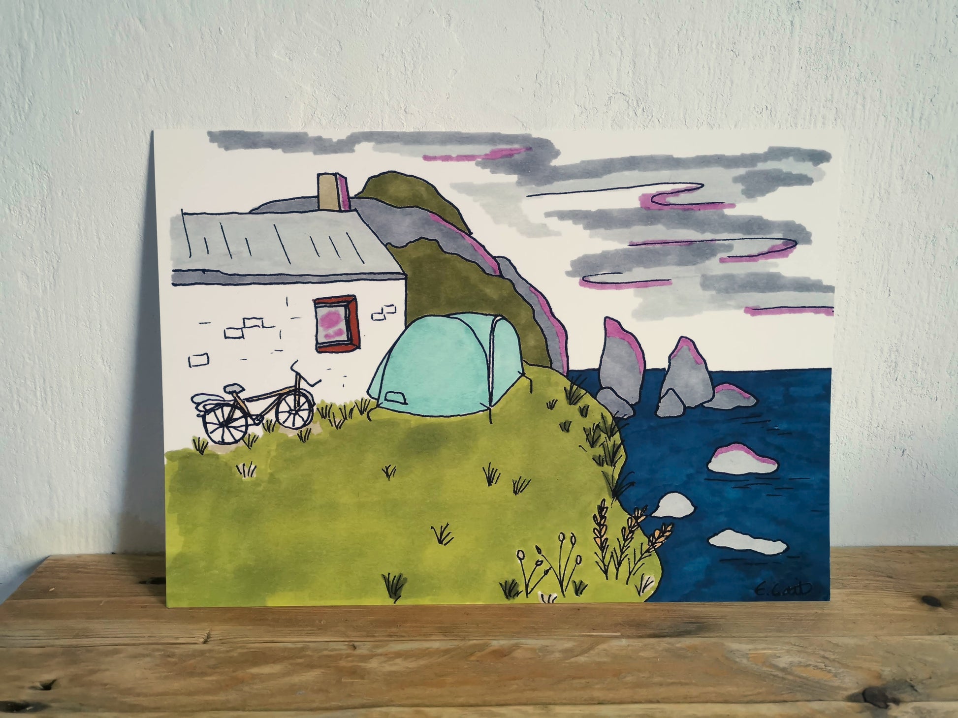 Hand-drawn landscape with a house, tent, and bicycle on a wooden surface.