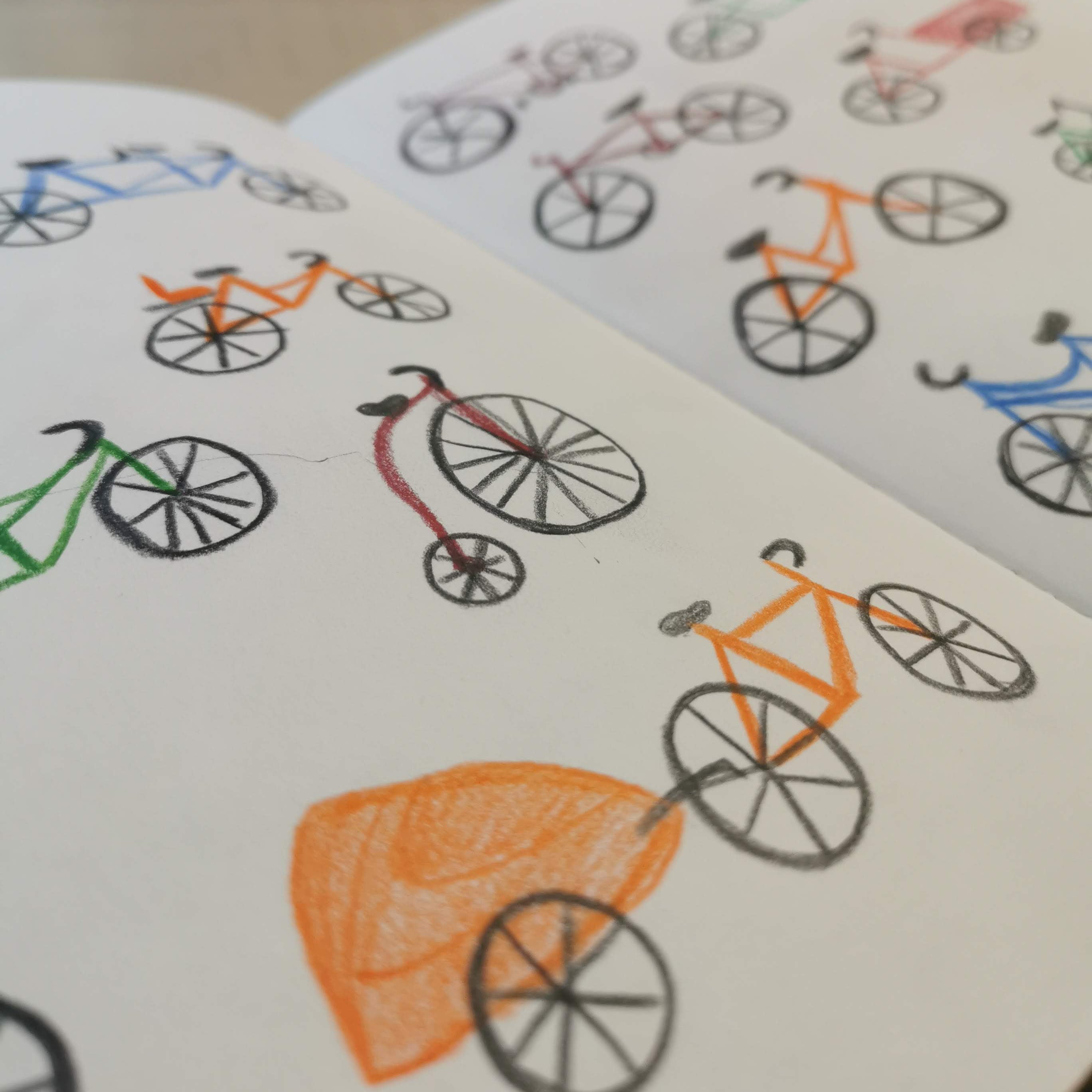 A close-up of an open notebook or book showing hand-drawn bicycle sketches on white pages, with one bicycle drawn in orange and black and another in green and black.